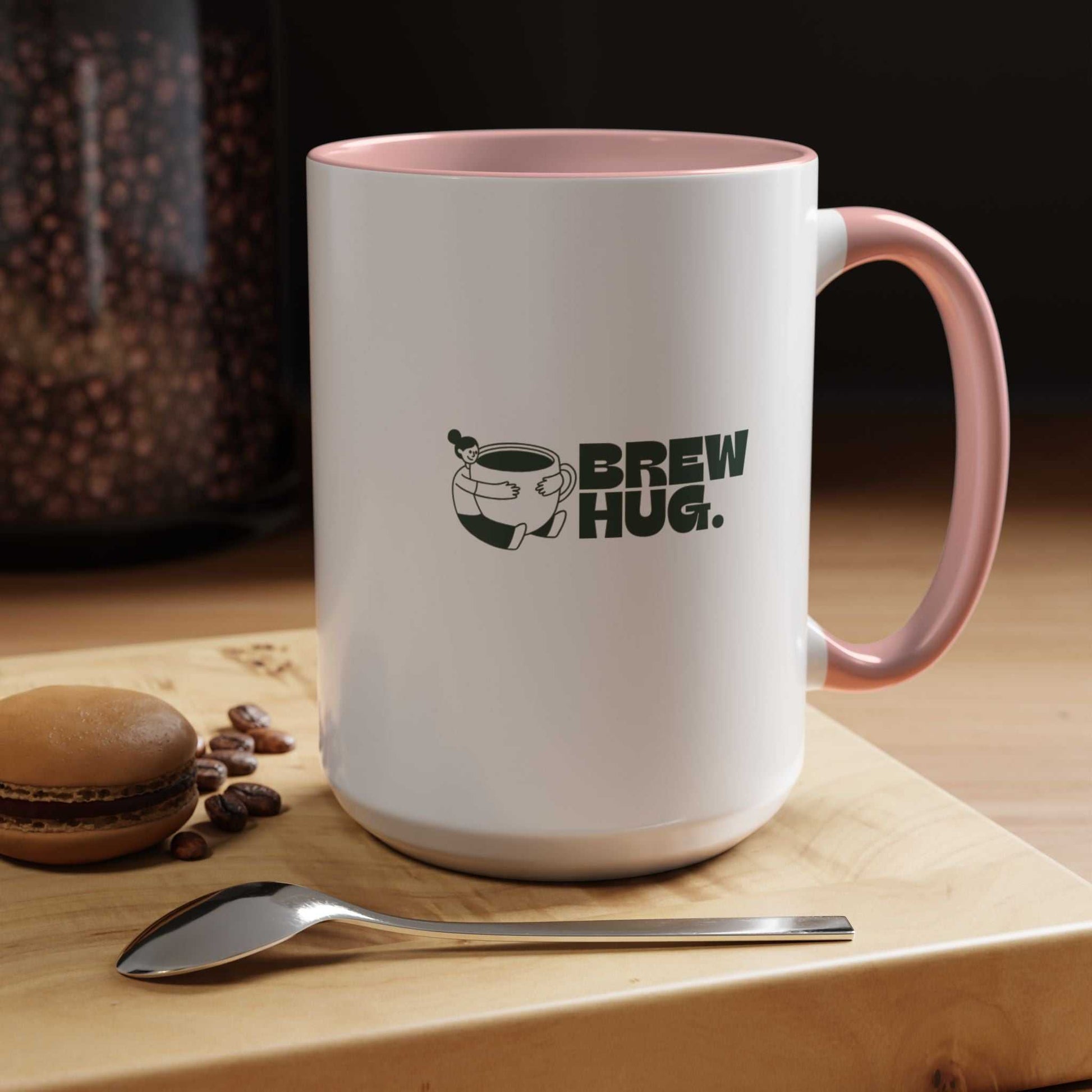 White mug with pink handle featuring 'Brew Hug' text on a wooden surface with coffee beans and a macaron.