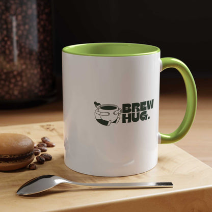 White mug with green interior and handle, featuring 'Brew Hug' text, on a wooden surface with coffee beans and a spoon.